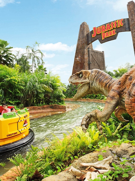 Visitors on Jurassic Park Rapids Adventure ride at Universal Studios Singapore, with dinosaur and jungle scenery.