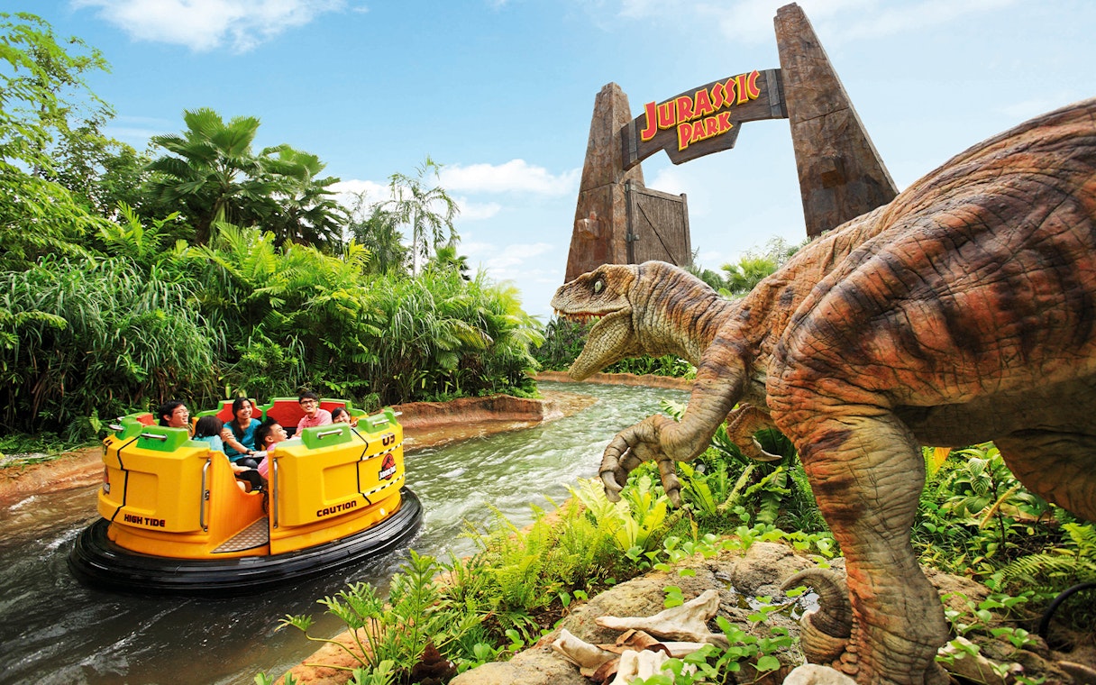 Visitors on Jurassic Park Rapids Adventure ride at Universal Studios Singapore, with dinosaur and jungle scenery.