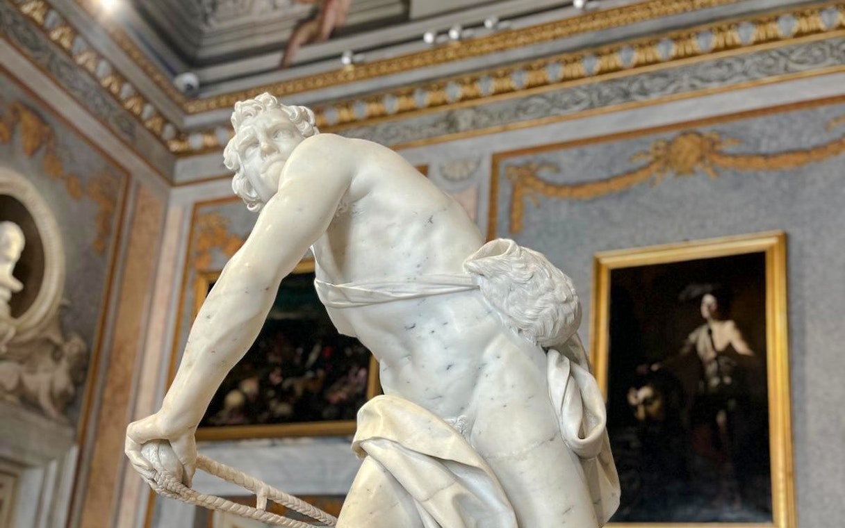 Marble statue in Borghese Gallery, Rome, with ornate paintings and decor in the background.