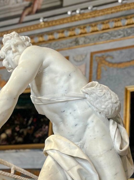 Marble statue in Borghese Gallery, Rome, with ornate paintings and decor in the background.