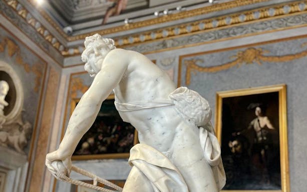 Marble statue in Borghese Gallery, Rome, with ornate paintings and decor in the background.
