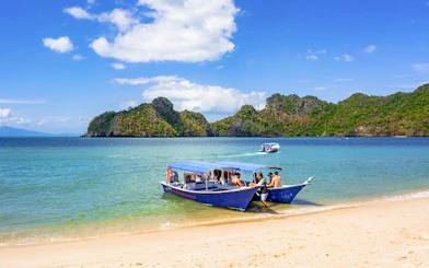 Boat docked on Langkawi beach with tourists, lush hills in background, part of Mangrove Discovery Adventure Tour.