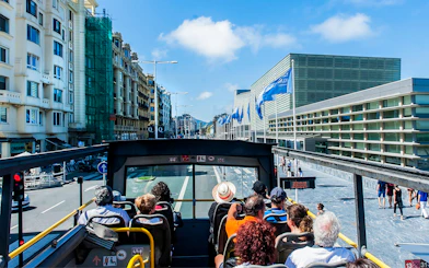 Open-top bus tour in San Sebastian city with passengers viewing modern architecture.