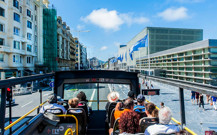 Open-top bus tour in San Sebastian city with passengers viewing modern architecture.