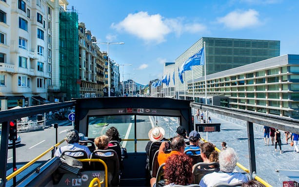 Open-top bus tour in San Sebastian city with passengers viewing modern architecture.
