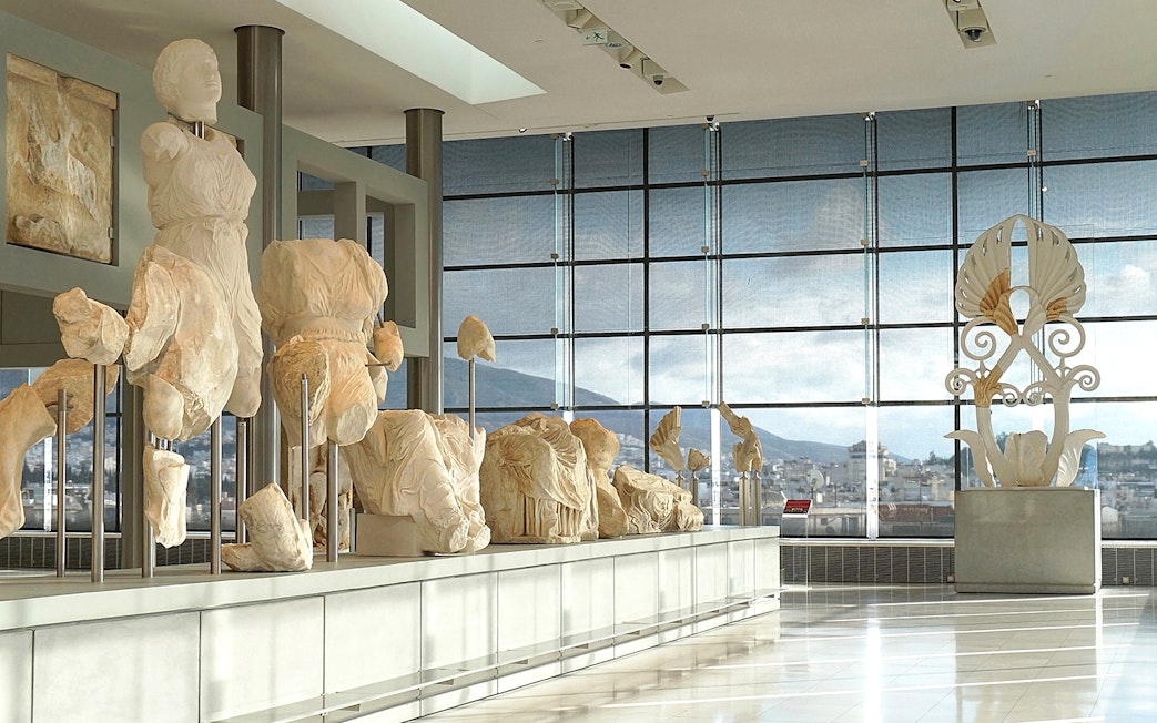 Ancient sculptures displayed in the Acropolis Museum with city view through large windows.