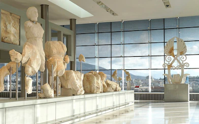 Ancient sculptures displayed in the Acropolis Museum with city view through large windows.