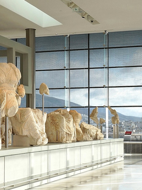 Ancient sculptures displayed in the Acropolis Museum with city view through large windows.