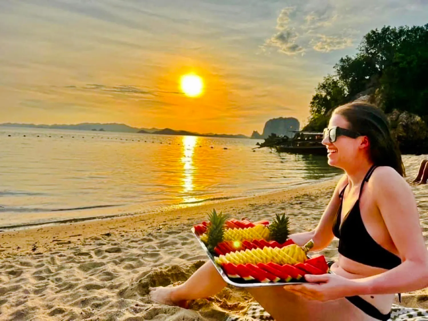 Sunset view on Krabi beach with a person enjoying a fruit platter, longtail boat in the background.