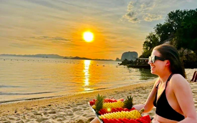 Sunset view on Krabi beach with a person enjoying a fruit platter, longtail boat in the background.