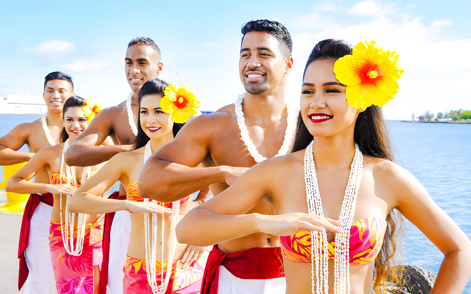 Hawaiian dancers in traditional attire with leis performing by the ocean.