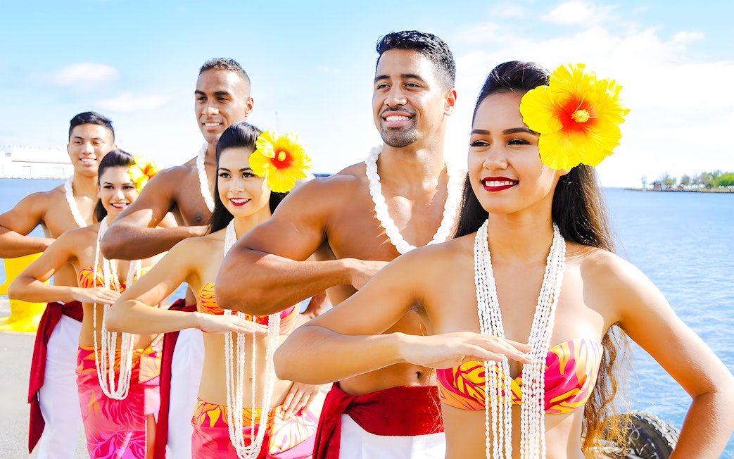 Hawaiian dancers in traditional attire with leis performing by the ocean.
