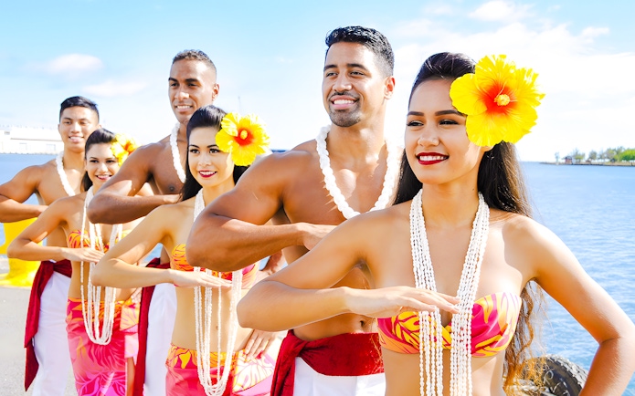 Hawaiian dancers in traditional attire with leis performing by the ocean.