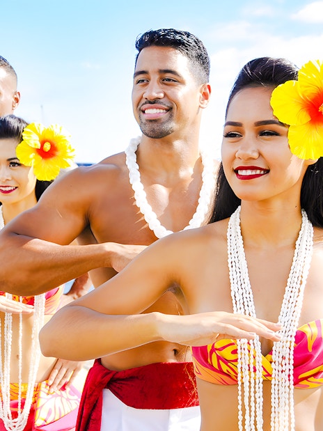 Hawaiian dancers in traditional attire with leis performing by the ocean.