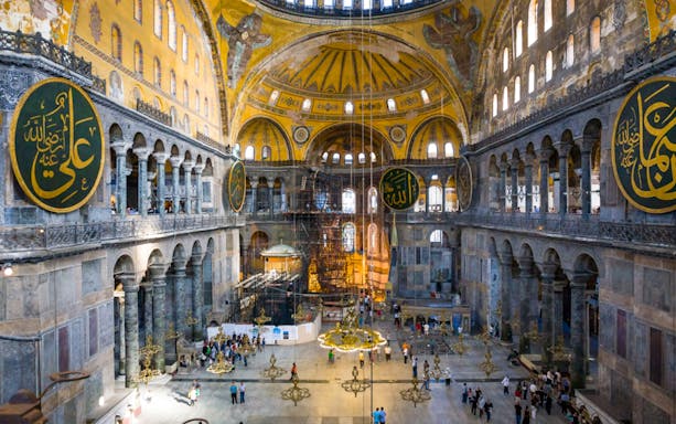 Interior of Hagia Sophia with visitors, showcasing ornate arches and large calligraphic medallions, Istanbul.