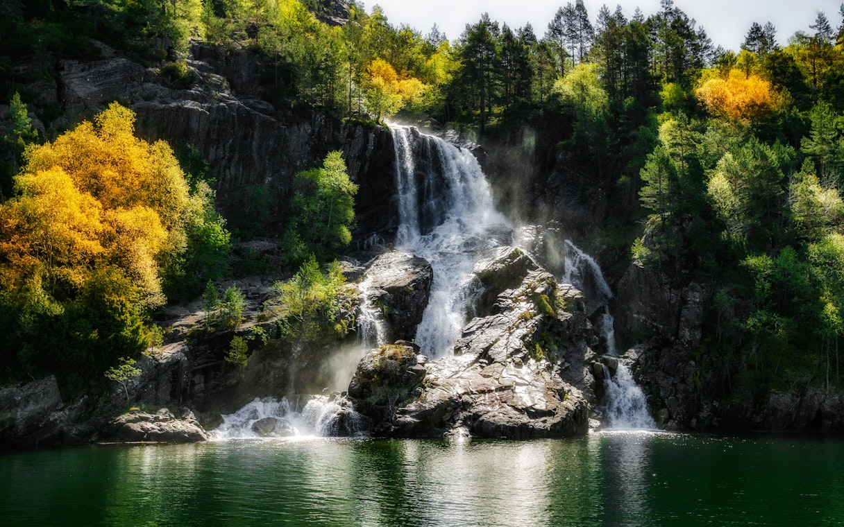Waterfall cascading over rocks in Lysefjord, Norway, surrounded by lush green and autumn trees.