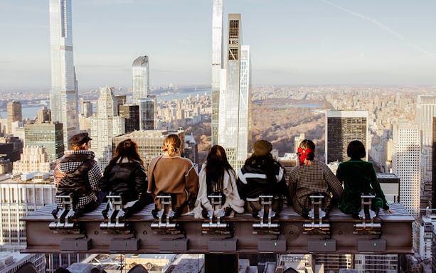 Visitors seated on a rooftop edge overlooking New York City skyline at Rockefeller Center.