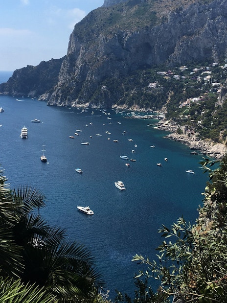 Boats on the blue waters of Capri Island with rocky cliffs and greenery, view from Naples day trip.