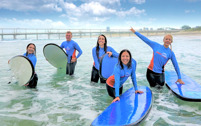 Beginner surfers in wetsuits holding boards in ocean near pier.