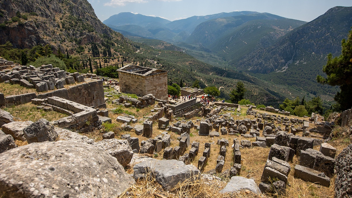Visitors exploring ancient ruins at Delphi archaeological site, Greece, with mountainous landscape.