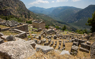 Visitors exploring ancient ruins at Delphi archaeological site, Greece, with mountainous landscape.