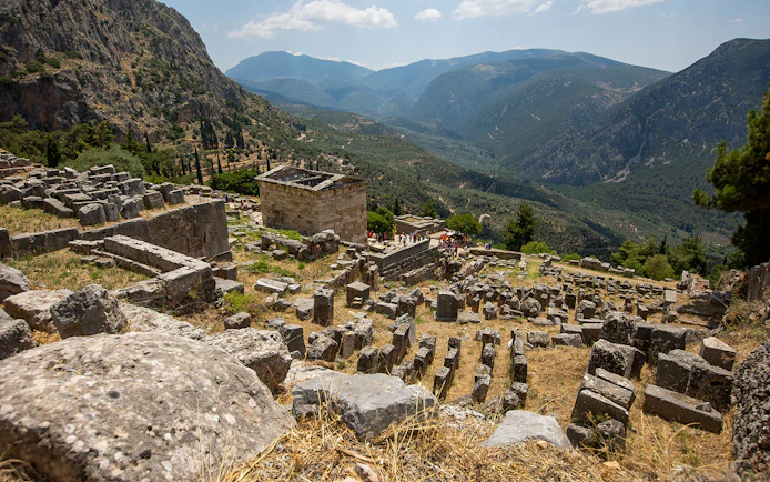 Visitors exploring ancient ruins at Delphi archaeological site, Greece, with mountainous landscape.