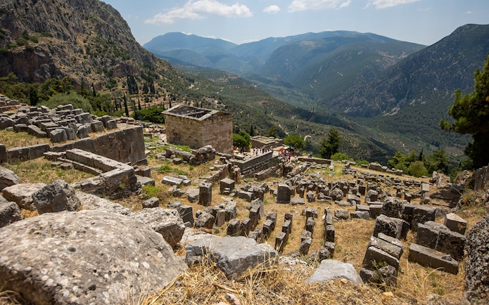 Visitors exploring ancient ruins at Delphi archaeological site, Greece, with mountainous landscape.