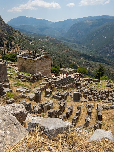 Visitors exploring ancient ruins at Delphi archaeological site, Greece, with mountainous landscape.