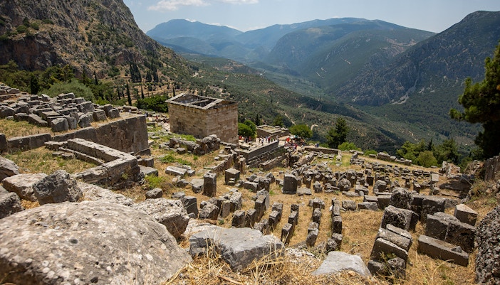 Visitors exploring ancient ruins at Delphi archaeological site, Greece, with mountainous landscape.
