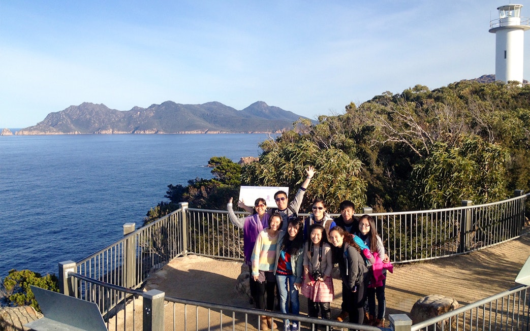 Group of tourists at Wineglass Bay lookout with ocean and lighthouse in background.