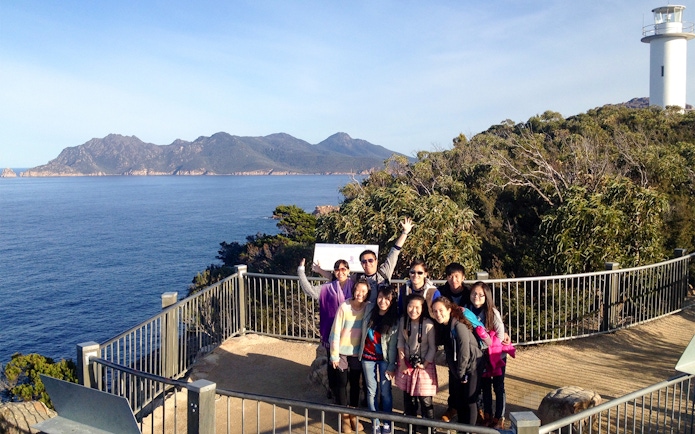 Group of tourists at Wineglass Bay lookout with ocean and lighthouse in background.