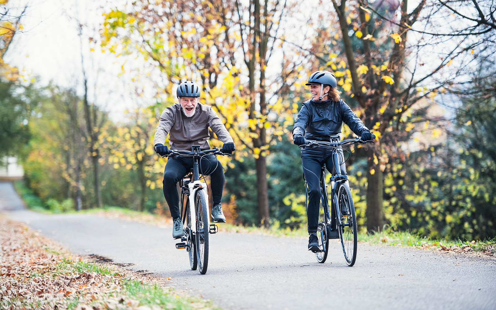Senior couple riding electrobikes on a tree-lined road in autumn.