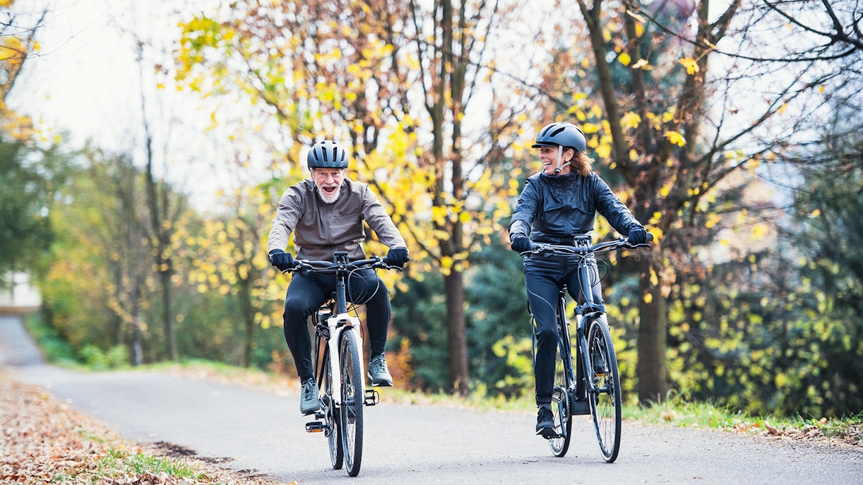 Senior couple riding electrobikes on a tree-lined road in autumn.