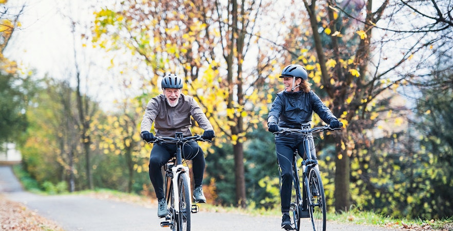 Senior couple riding electrobikes on a tree-lined road in autumn.