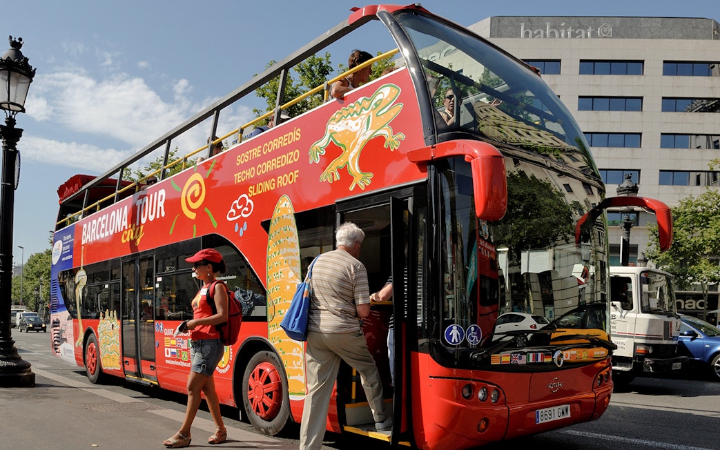 Barcelona tour bus with passengers boarding near city landmarks.