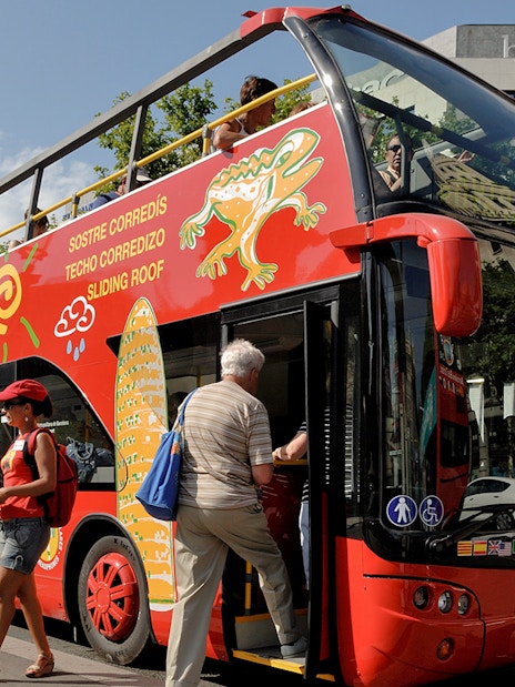 Barcelona tour bus with passengers boarding near city landmarks.