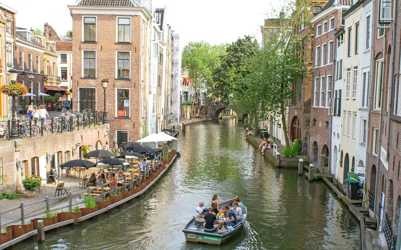 Boat on Utrecht canal with people dining at waterside cafes, showcasing historic buildings and greenery.