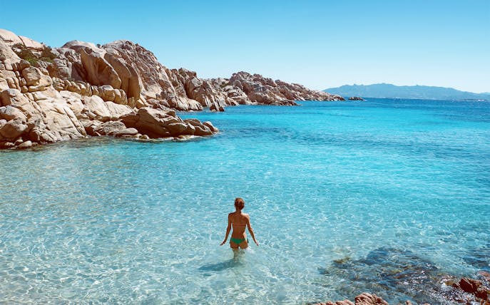 Person wading in clear waters of Gulf of Orosei, rocky coastline in background.