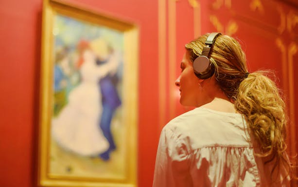 Visitor with headphones viewing French Impressionist painting at NGV Melbourne exhibition.