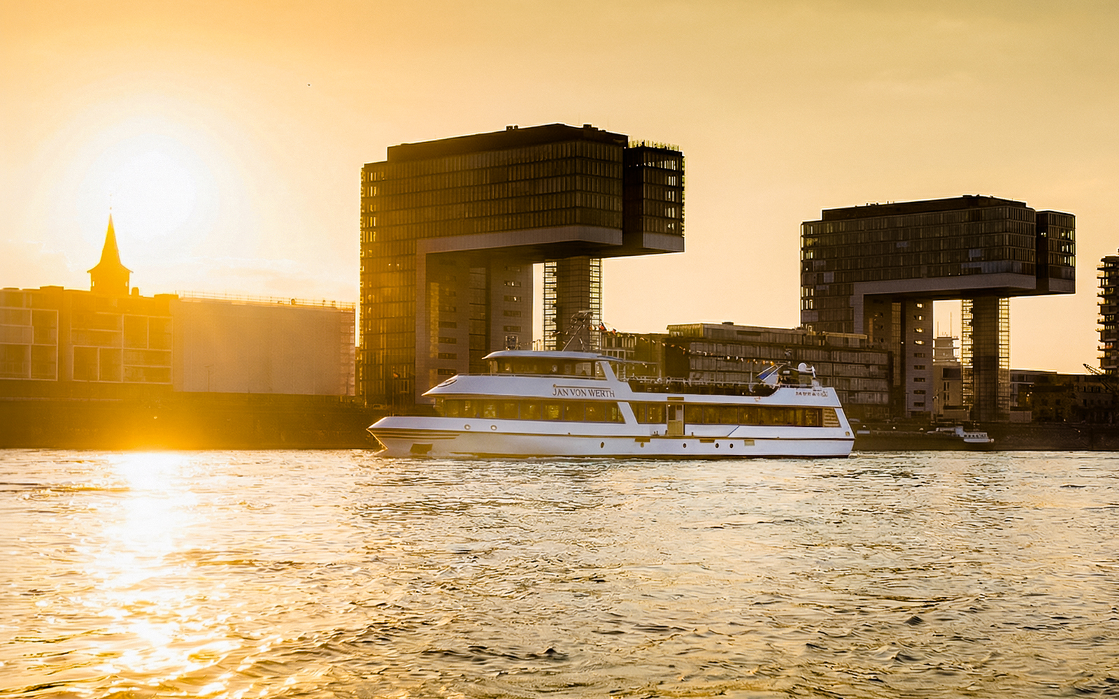 Cruise boat on Rhine River at sunset with Cologne's crane houses in view.
