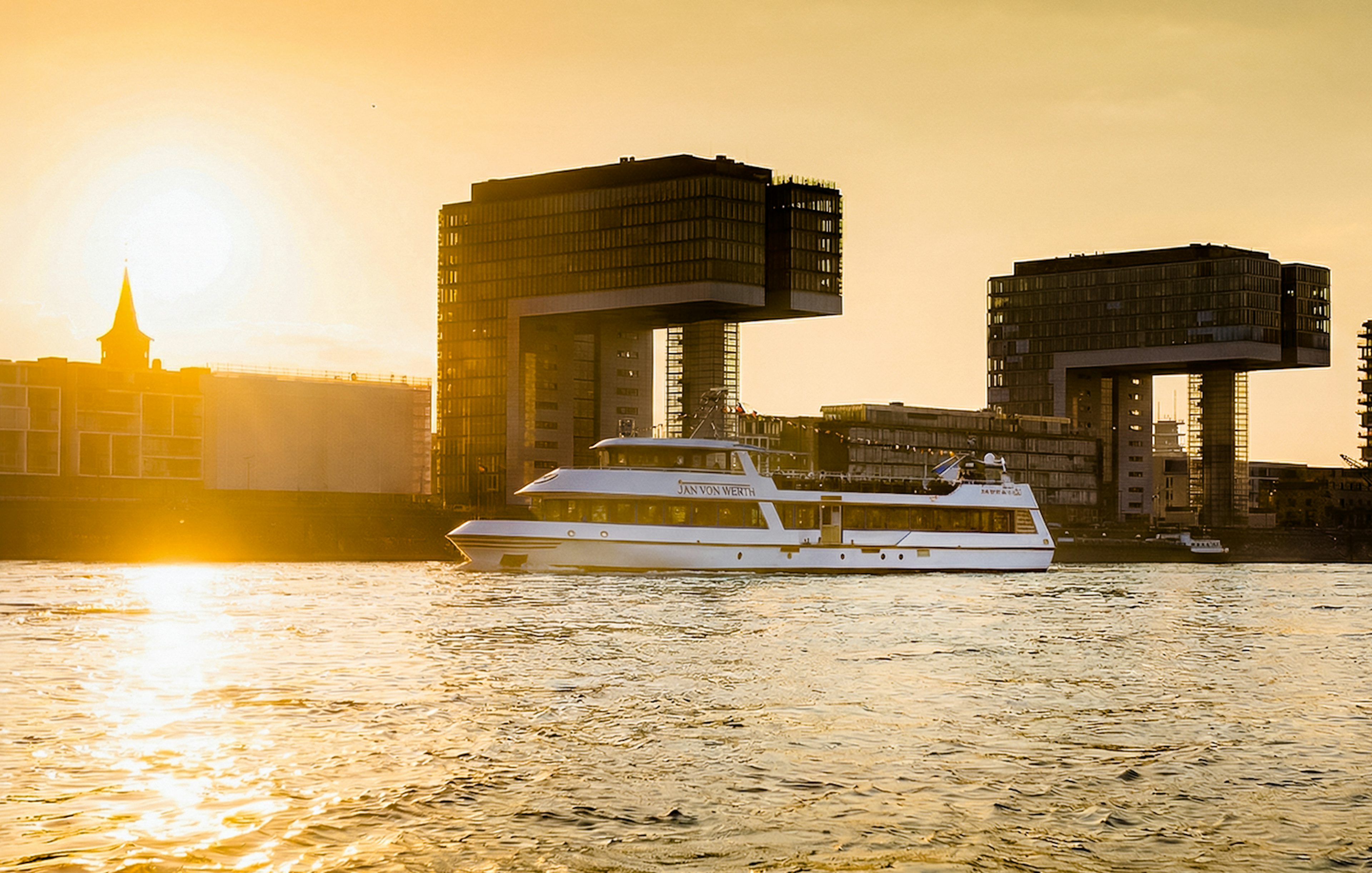 Cruise boat on Rhine River at sunset with Cologne's crane houses in view.