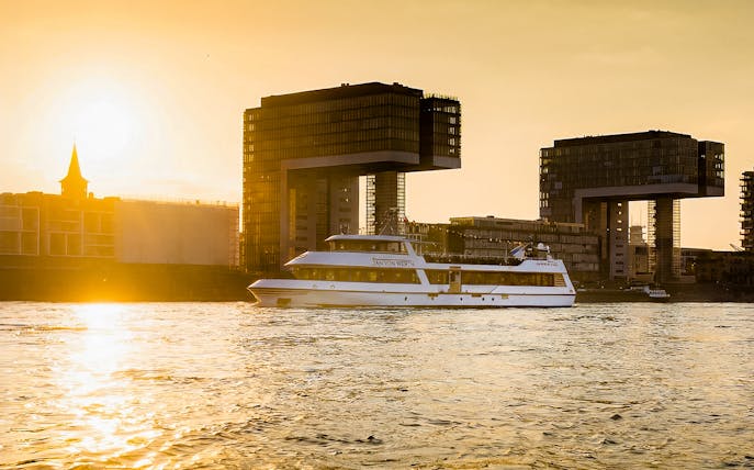 Cruise boat on Rhine River at sunset with Cologne's crane houses in view.