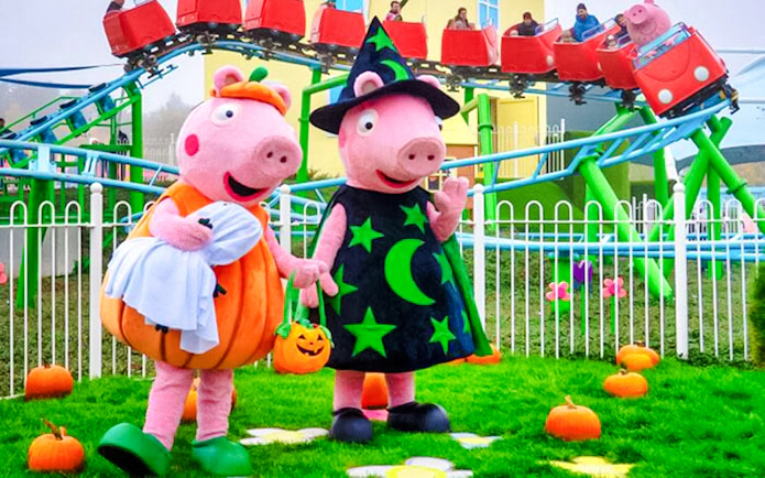 Peppa Pig characters in Halloween costumes at Peppa Pig Park with roller coaster in background.
