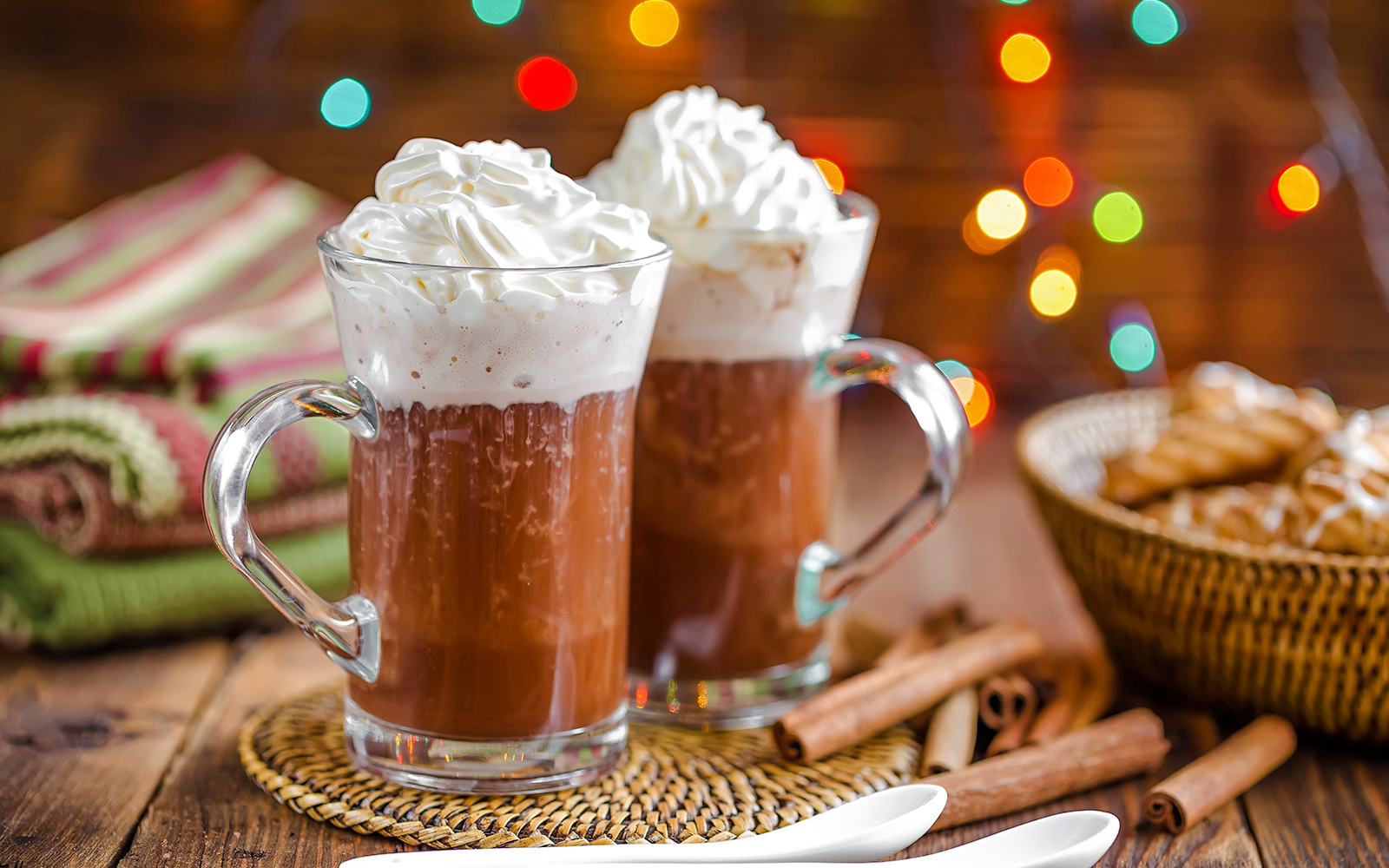 Hot chocolate with whipped cream in glass mugs, Christmas lights in background.
