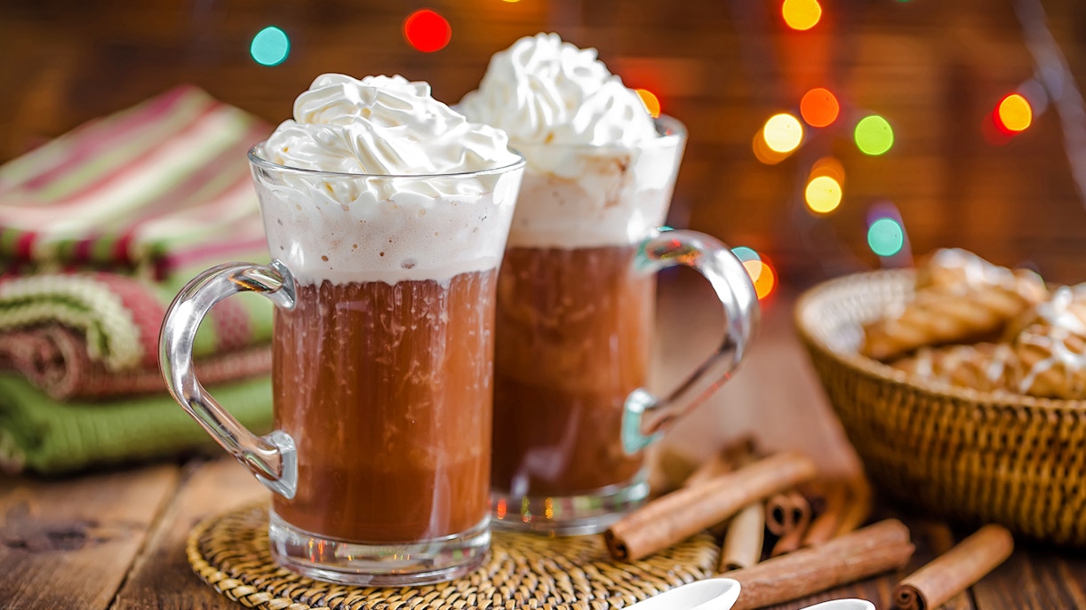 Hot chocolate with whipped cream in glass mugs, Christmas lights in background.