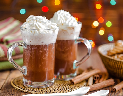 Hot chocolate with whipped cream in glass mugs, Christmas lights in background.
