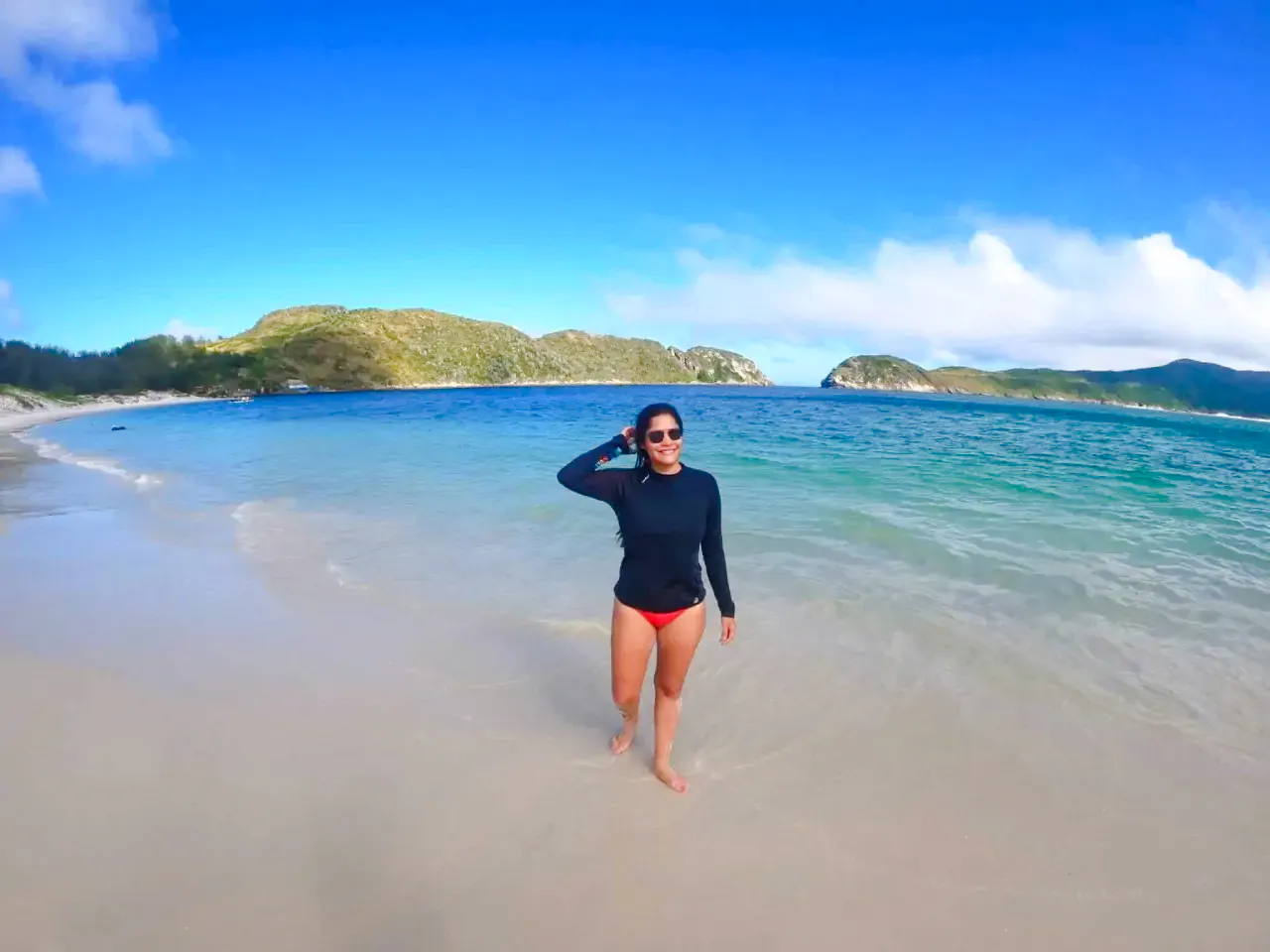 Person walking on a beach with clear blue water and distant hills in Arraial do Cabo, Brazil.