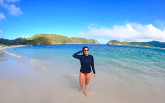 Person walking on a beach with clear blue water and distant hills in Arraial do Cabo, Brazil.