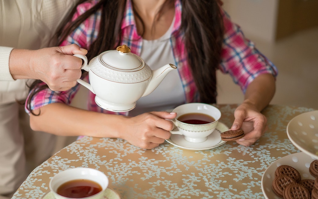 Pouring tea from a porcelain teapot into a cup on a patterned tablecloth.