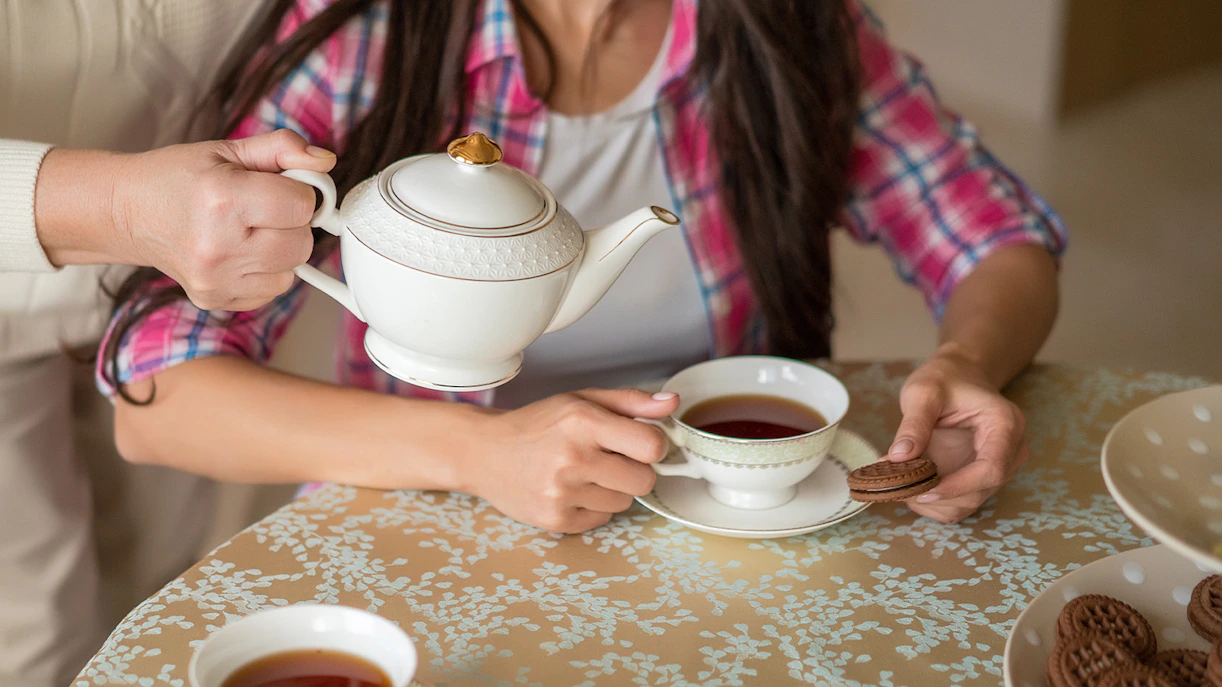 Pouring tea from a porcelain teapot into a cup on a patterned tablecloth.